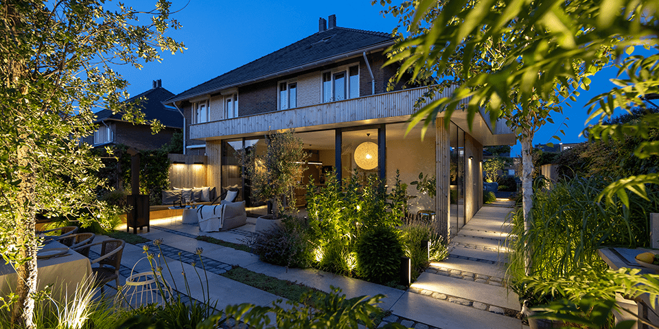 In einem beleuchteten Garten mit Terrasse sind unterschiedliche Formen direkter und indirekter Gartenbeleuchtung zu sehen: Au&szlig;enwandleuchten, Outdoor-H&auml;ngeleuchten, LED-Streifen, Wegeleuchten, Bodeneinbauleuchten und Gartenstrahler. 