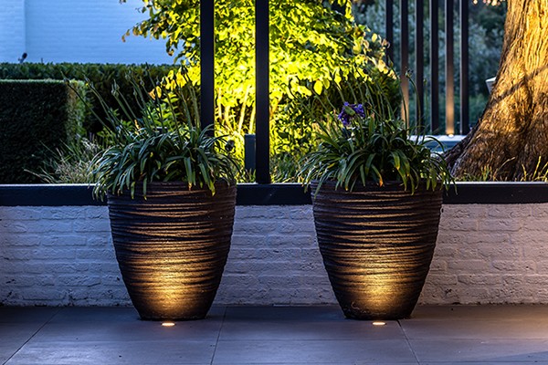 Bodenleuchten auf einer Terrasse werfen ein dekoratives Licht auf zwei Pflanzk&uuml;bel. Im Hintergrund ist ein beleuchteter Garten zu sehen.