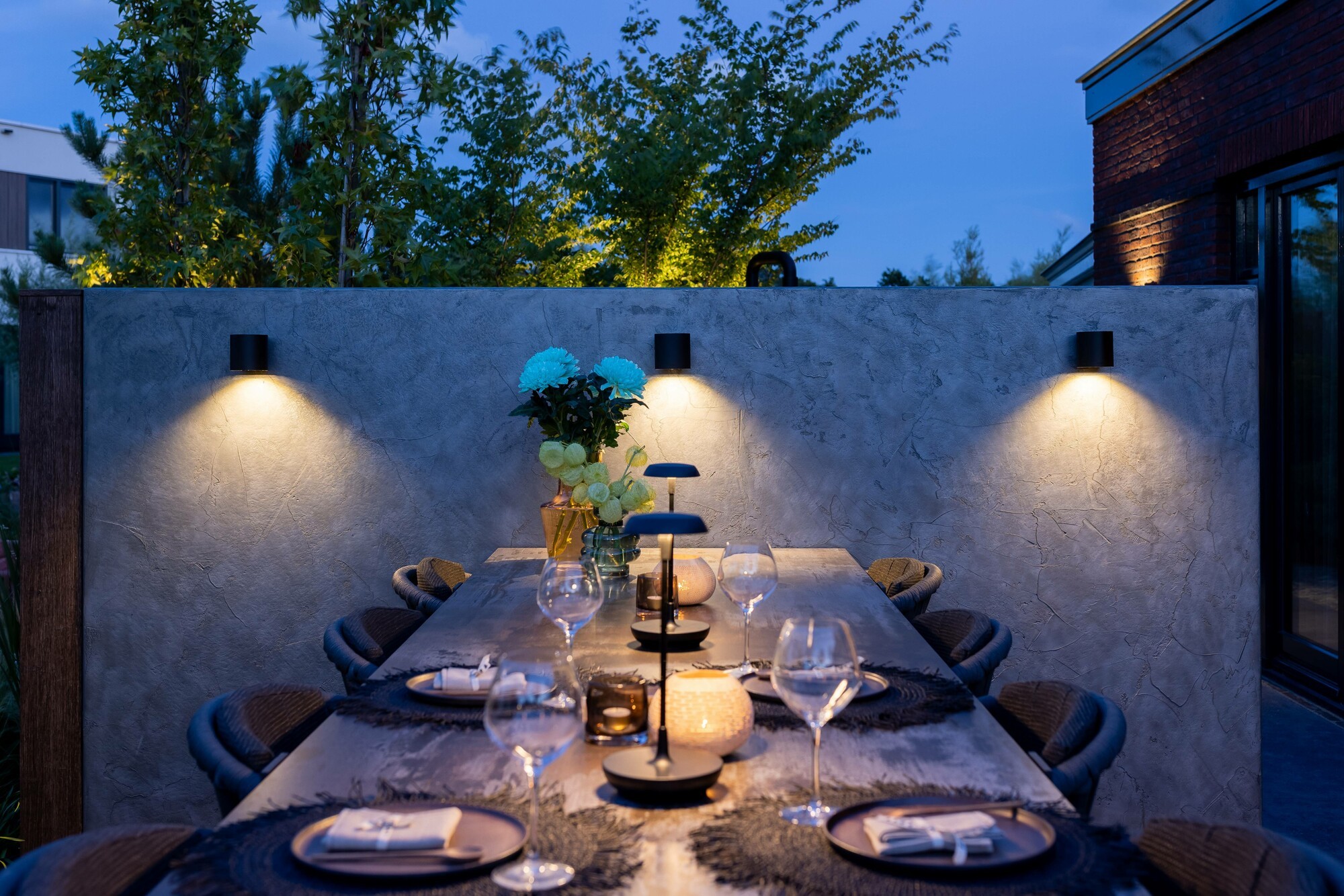 Eine Mauer mit drei LED-Au&szlig;enwandleuchten, die eine angenehme Beleuchtung erzeugen. Davor ein gedeckter Gartentisch, der indirekt von den Au&szlig;enwandleuchten und direkt von Tischleuchten erhellt wird. 
