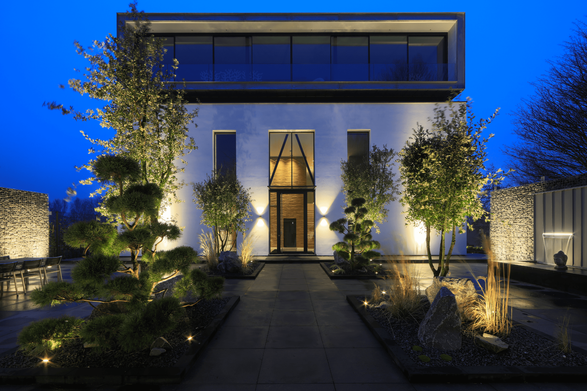 Ein Haus mit Fassadenbeleuchtung, die au&szlig;en in der Abendd&auml;mmerung ein stimmungsvolles Licht verbreitet. Davor liegt ein moderner Garten mit dekorativer Baumbeleuchtung.