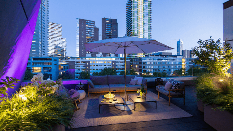 Eine moderne Dachterrasse in einer Gro&szlig;stadt wird mithilfe von exklusiver smarter in-lite-Au&szlig;enbeleuchtung und intelligentem System-Zubeh&ouml;r in farbiges Licht getaucht. Zu sehen sind eine beleuchtete Lounge-Ecke mit Sonnenschirm, mehrere beleuchtete Gr&uuml;npf