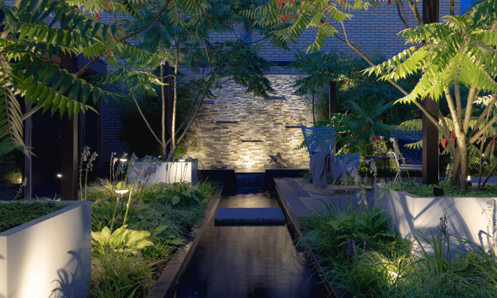 Ein stilvoller Au&szlig;enbereich mit modernem Gartenteich wird von unterschiedlichen Au&szlig;enleuchten in stimmungsvolles Licht getaucht. Zu sehen sind Stehleuchten, Gartenstrahler und Teichbeleuchtung. 